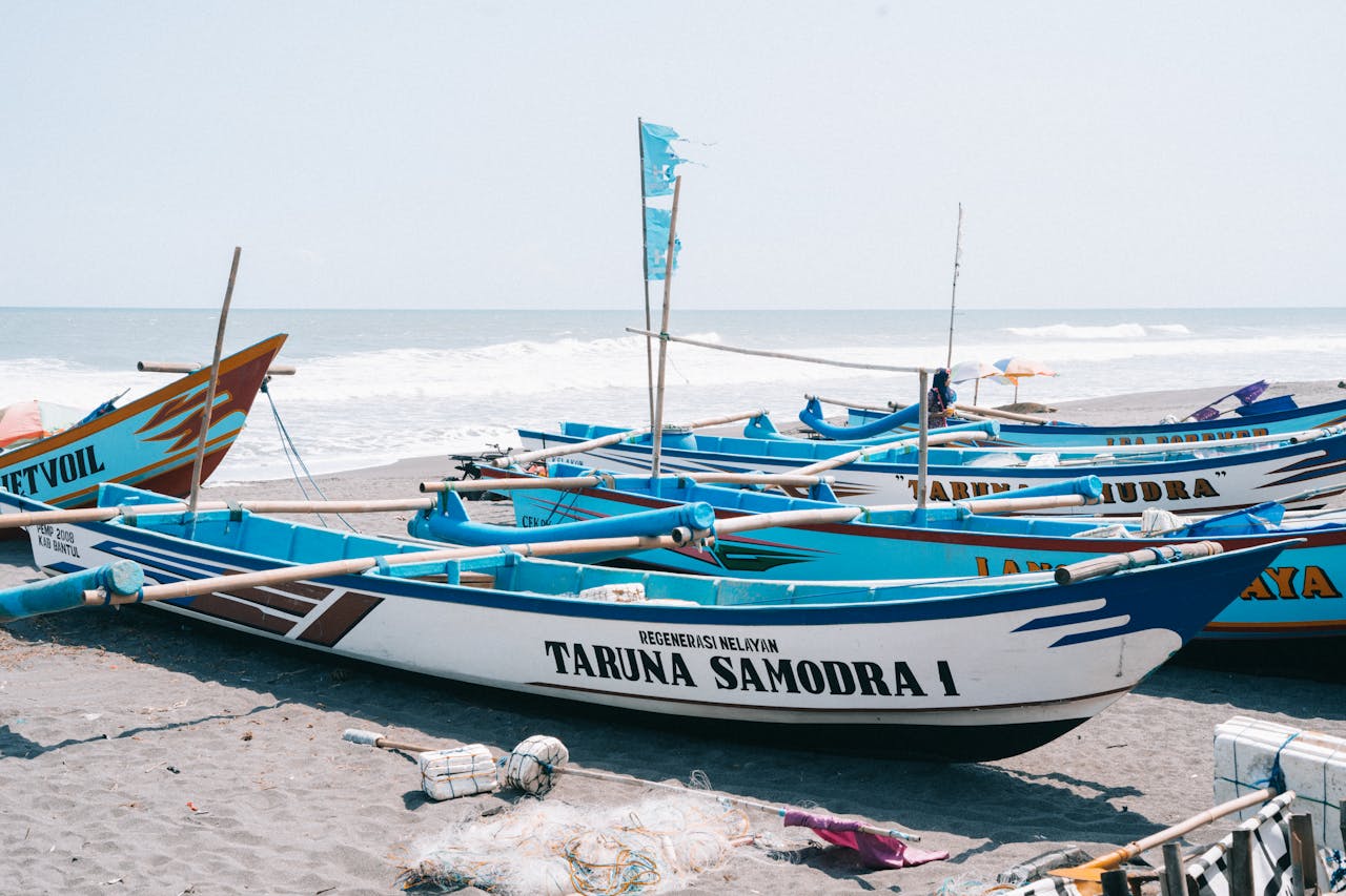 who-we-are Rows of vibrant fishing boats rest on the beach with waves in the background.